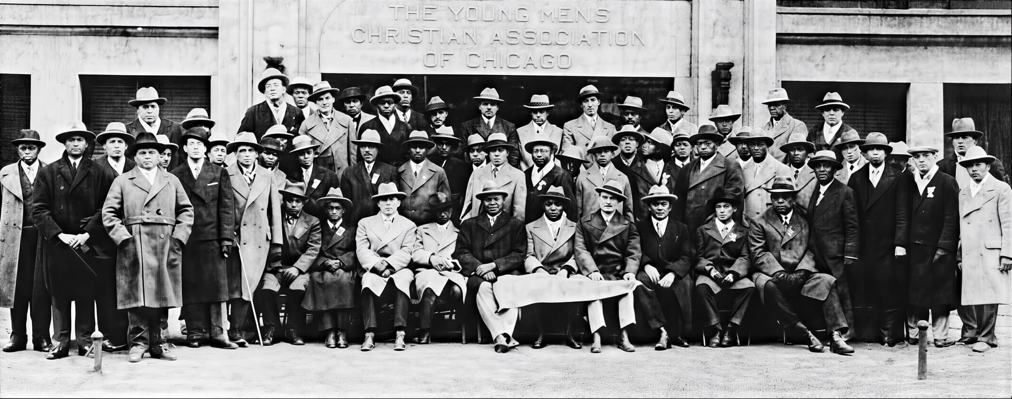 A large group of Omega men in overcoats and hats, seated and standing for a panoramic portrait beneath the carved stone facade of the Young Men's Christian Association of Chicago